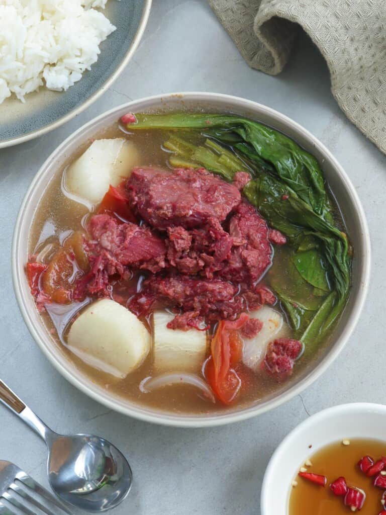 sinigang na corned beef in a white bowl with a plate of rice on the side