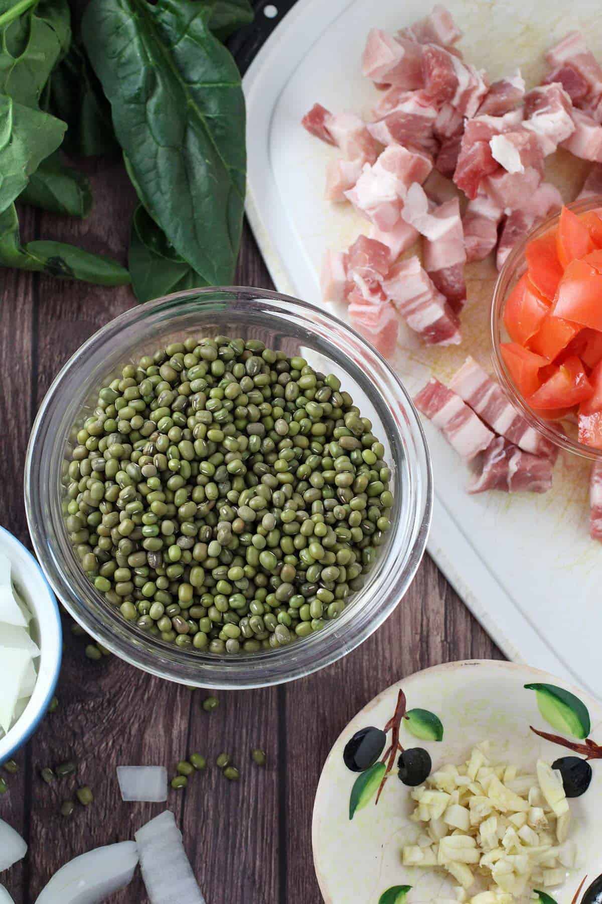 dry mung beans, diced raw pork belly, chopped tomatoes, spinach leaves, chopped onions, coconut milk on a wooden board