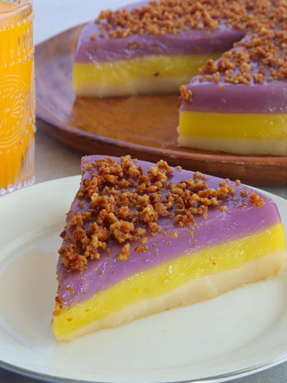 slice of Sapin-Sapin on a white plate with a whole piece on a wooden plate and glass of orange juice in the background.