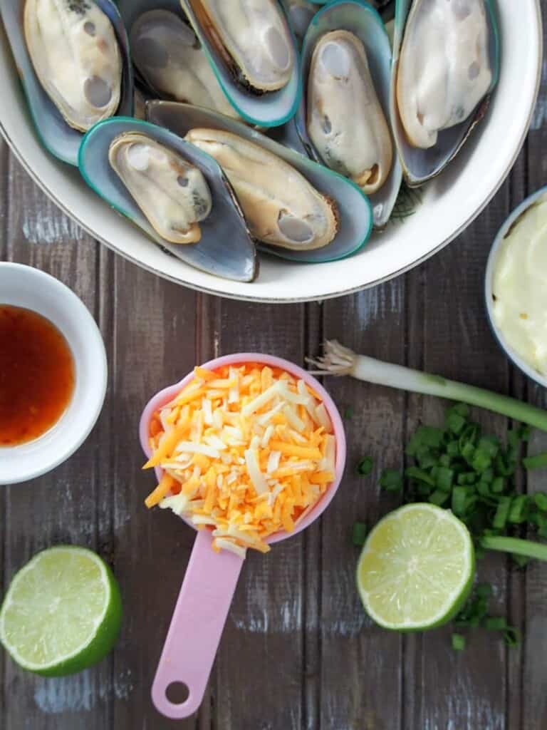 half-shell mussels in a white bowl, shredded cheese, mayonnaise, sweet chili sauce, limes