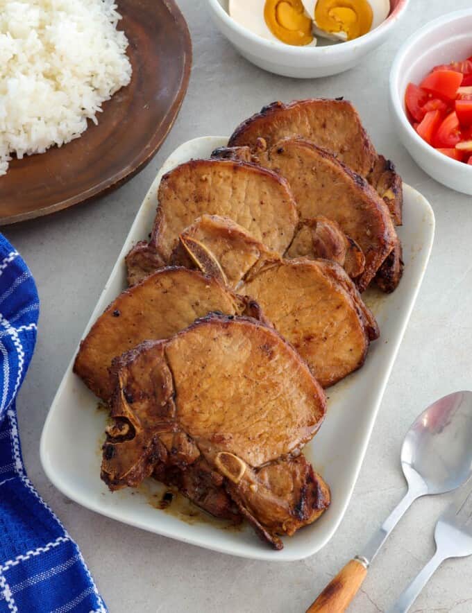 Toyomansi Pork Chops on a white platter with steamed rice, chopped tomatoes, and salted eggs on the side