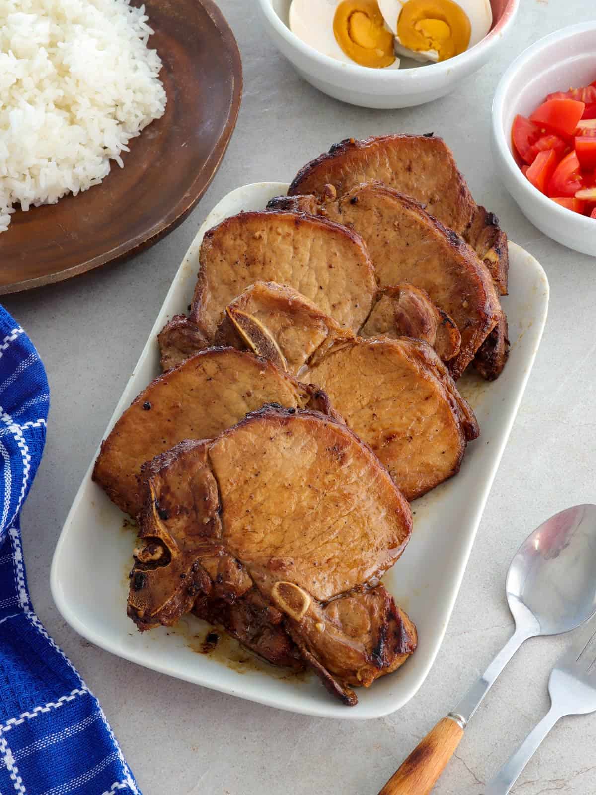 Toyomansi Pork Chops on a white platter with steamed rice, chopped tomatoes, and salted eggs on the side