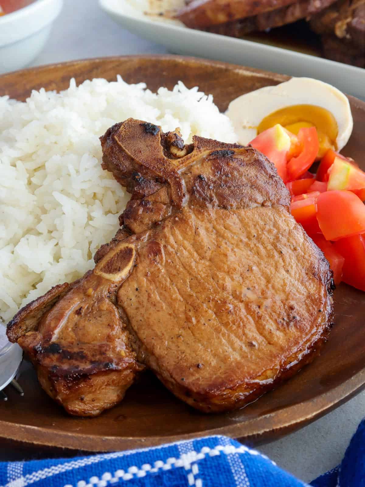 Toyomansi Pork Chops on a wooden plate with steamed rice, chopped tomatoes, and salted eggs on the side