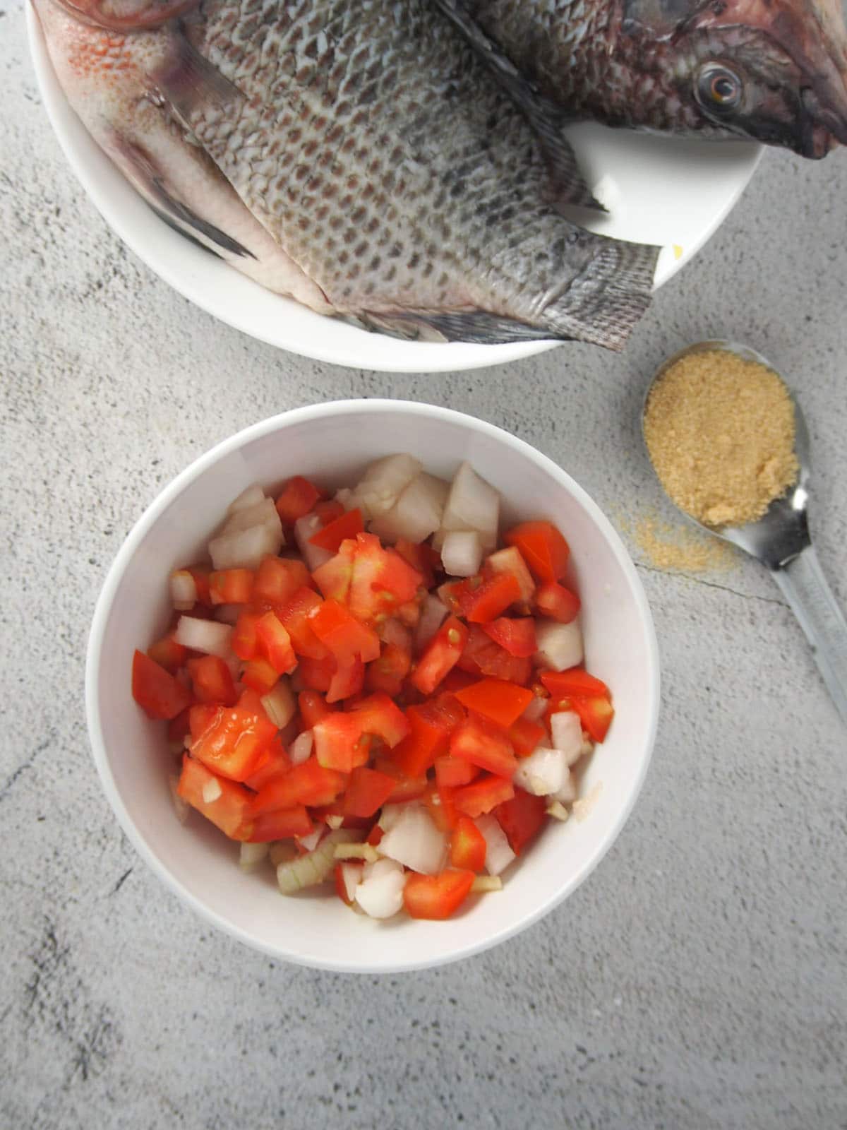 a bowl of chopped tomatoes, onions, garlic, and ginger and a plate of raw tilapia fish on the side