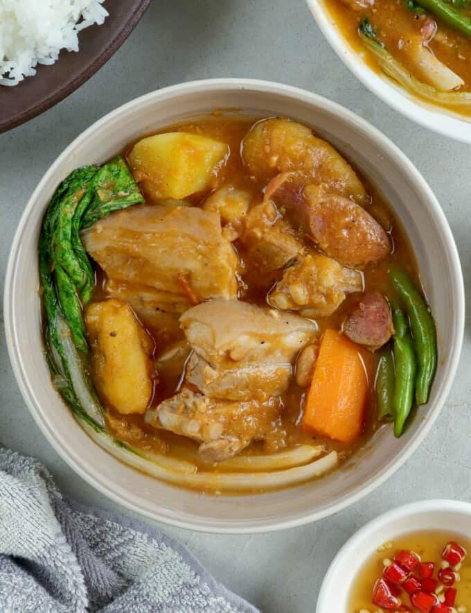 pocherong baboy with pork and beans, banana, potatoes, carrots and pechay in a white bowl with a plate of steamed rice on the side