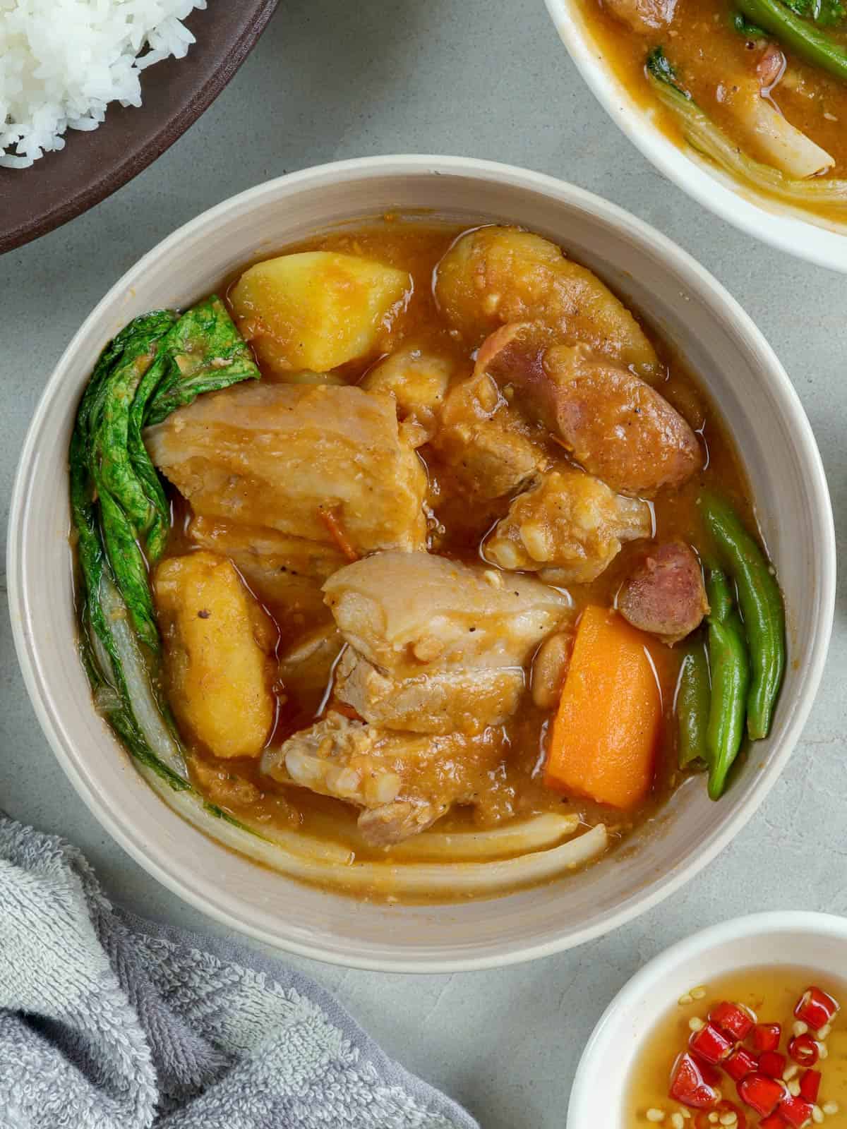 pocherong baboy with pork and beans, banana, potatoes, carrots and pechay in a white bowl with a plate of steamed rice on the side