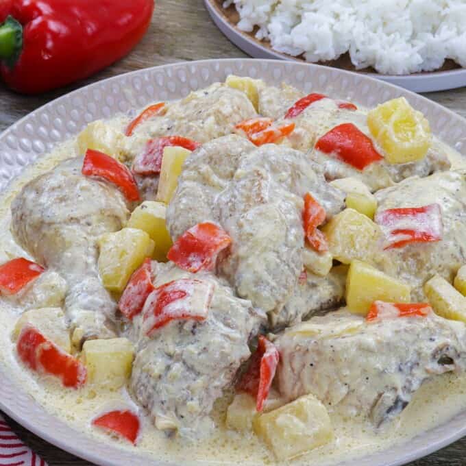 Pininyahang Manok on a white serving platter with a plate of steamed rice and red bell peppers in the background.