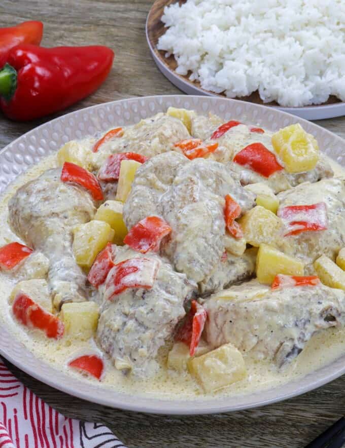 Pininyahang Manok on a white serving platter with a plate of steamed rice and red bell peppers in the background.