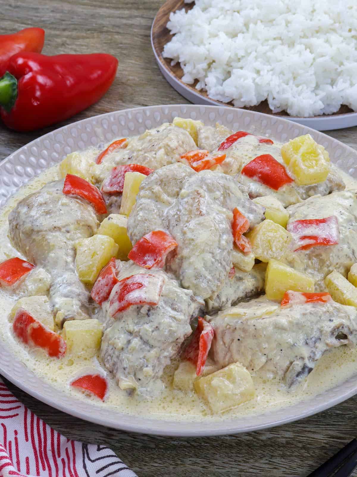 Pininyahang Manok on a white serving platter with a plate of steamed rice and red bell peppers in the background.