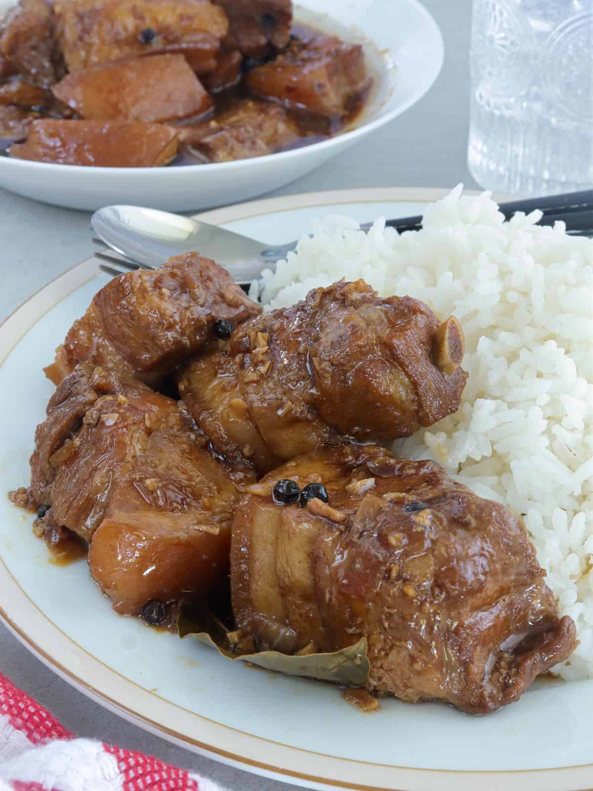 pork adobo with oyster sauce on a white serving plate with steamed rice and glass of water and a bowl of adobo in the background.