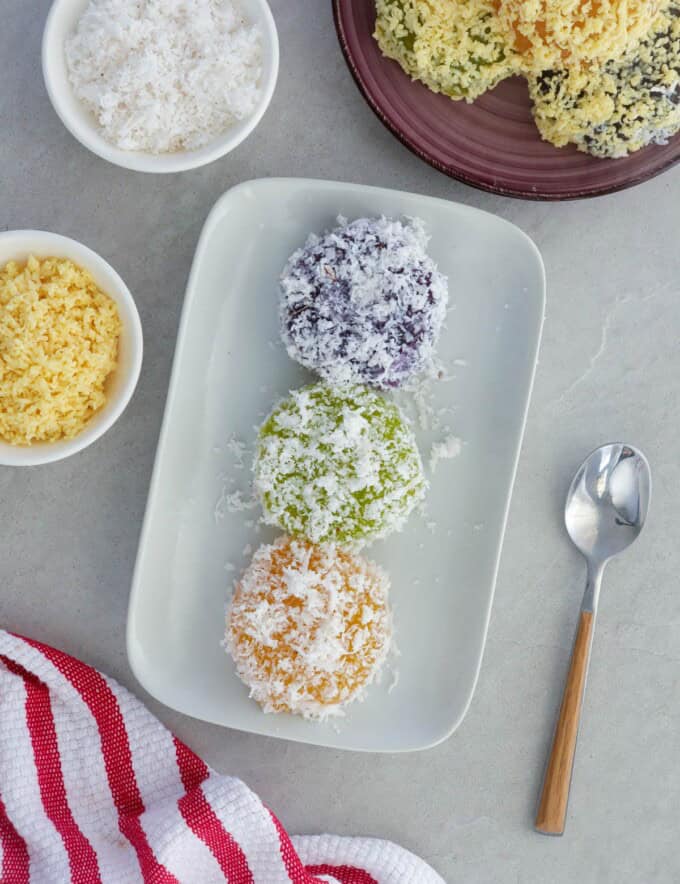 pichi-pichi with different flavors and coated with grated coconut on a white platter with a bowl of shredded cheese in the background.