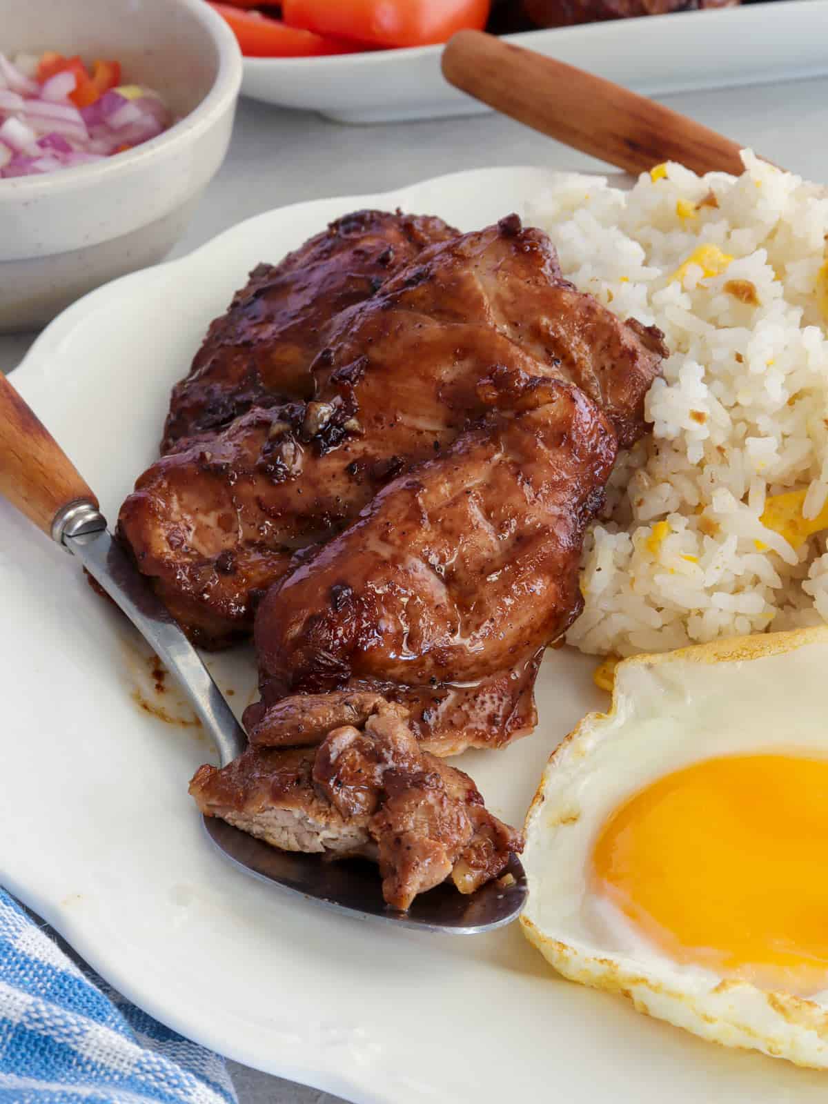 Chicken Tapa with garlic fried rice and sunny side up eggs on a white plate with a bowl of chopped tomatoes on the side.