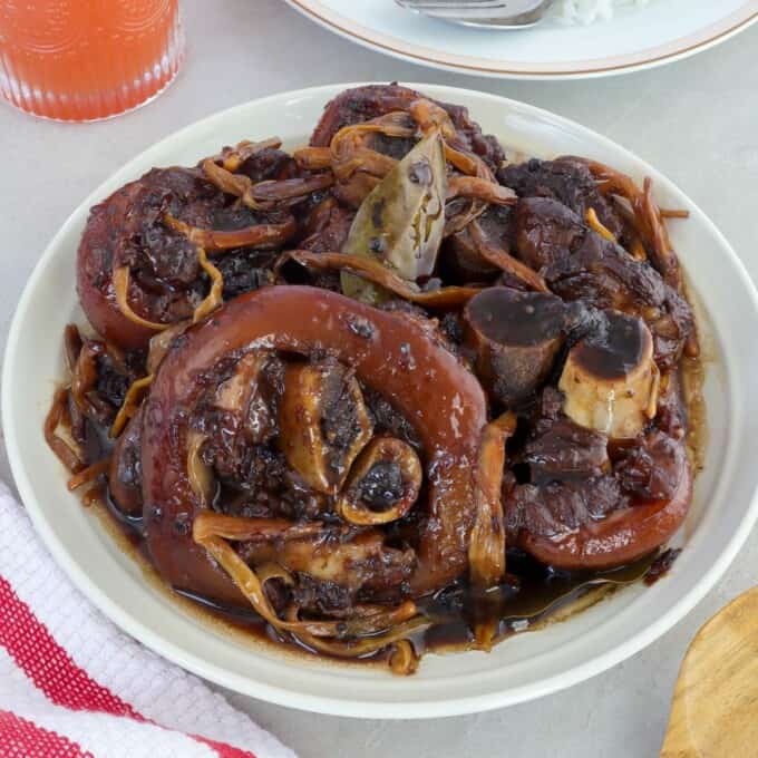 paksiw na pata in a white serving bowl with a a plate of steamed rice on the side