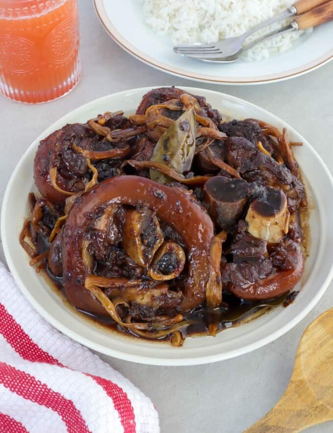 paksiw na pata in a white serving bowl with a a plate of steamed rice on the side