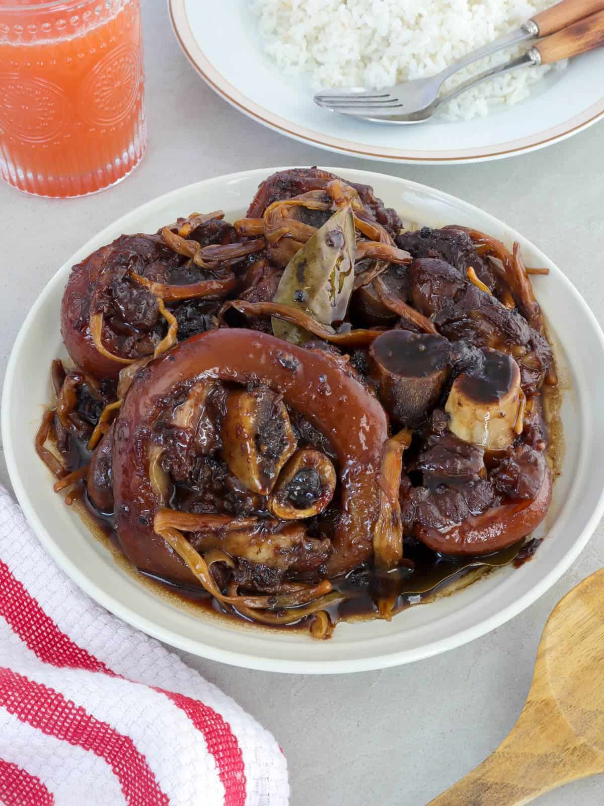 paksiw na pata in a white serving bowl with a a plate of steamed rice on the side