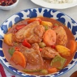 kalderatang manok in a blue serving bowl with a plate of steamed rice in the background.