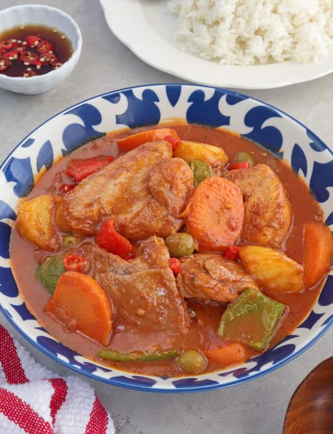 kalderatang manok in a blue serving bowl with a plate of steamed rice in the background.