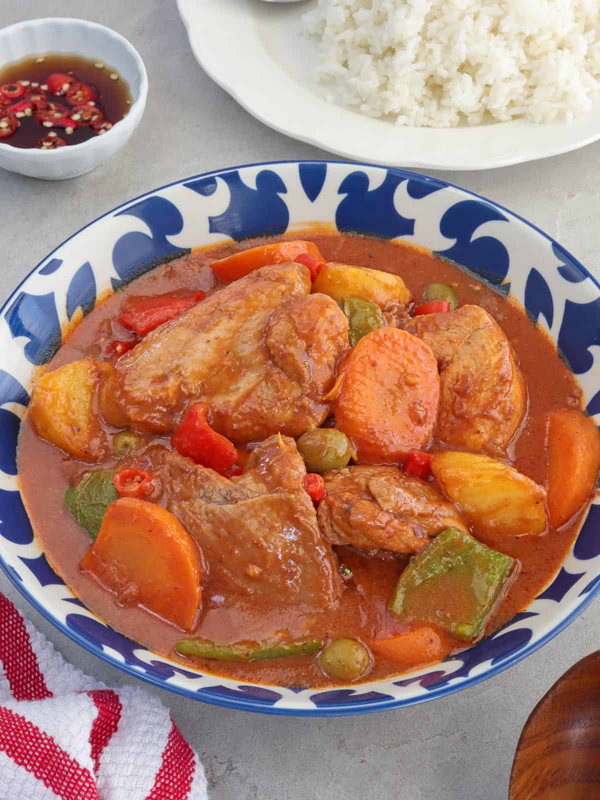 kalderatang manok in a blue serving bowl with a plate of steamed rice in the background.