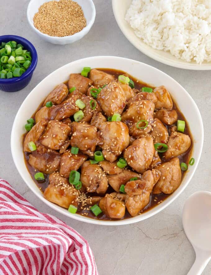 Sesame Chicken in a bowl with a plate of steamed rice in the background.