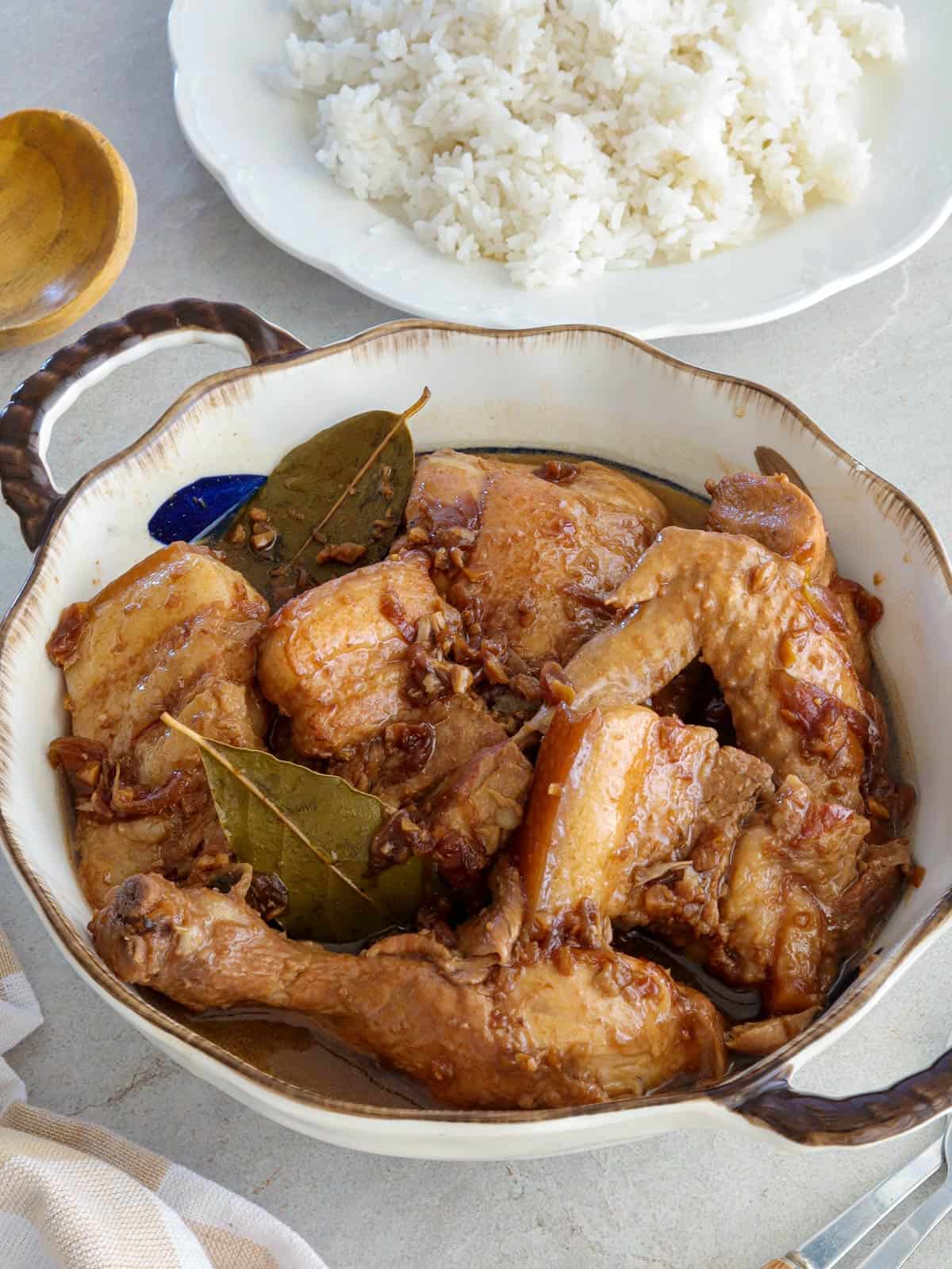 Chicken and Pork Adobo in a serving bowl with a plate of steamed rice in the background.