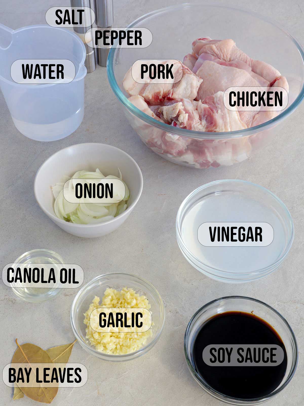 pork belly cubes, cut up chicken, garlic, soy sauce, vinegar, bay leaves, oil, onions in bowls and salt and pepper shakers in the background.