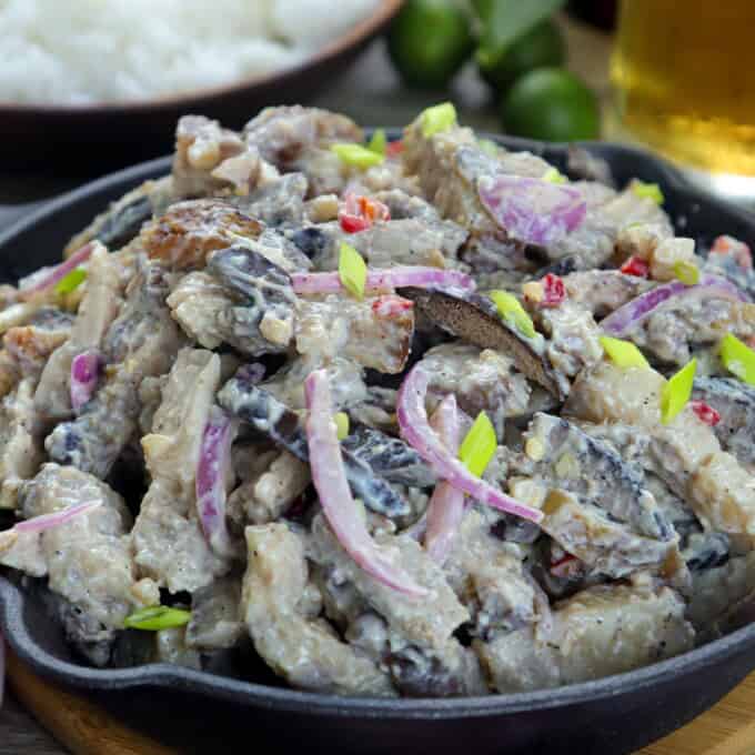 Dinakdakan in a cast iron skillet with a bowl of steamed rice on the side and a glass of beer.