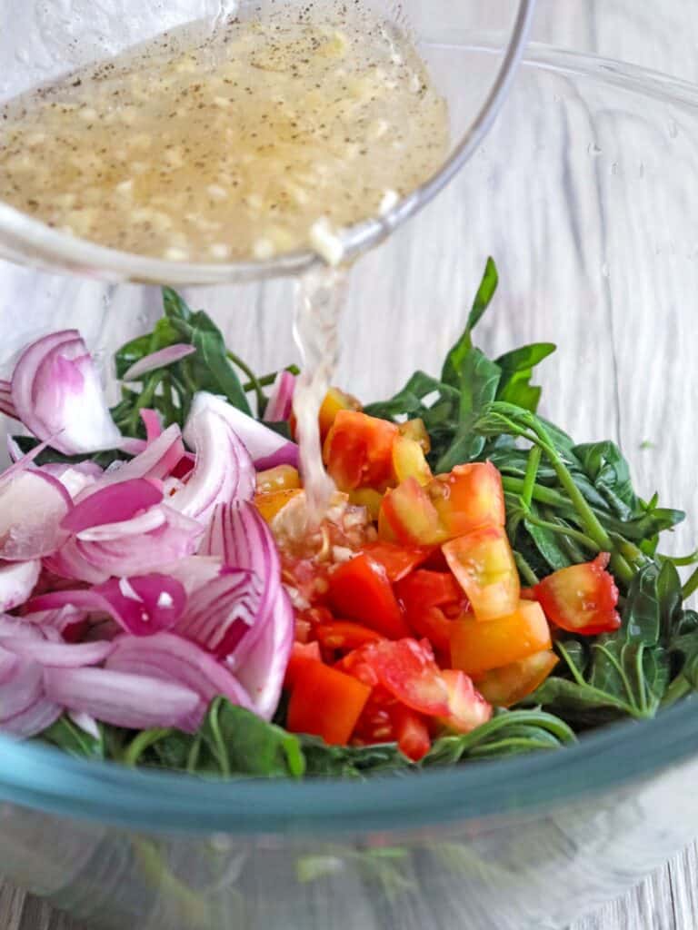 pouring vinaigrette dressing over kamote tops salad with tomatoes and red onions in a glass bowl