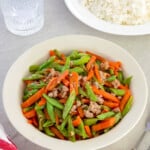 green bean stir-fry with carrots and minced pork on a white serving platter with a plate of steamed rice in the background.
