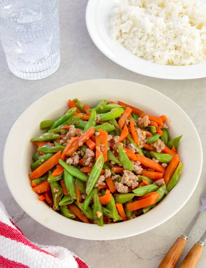 green bean stir-fry with carrots and minced pork on a white serving platter with a plate of steamed rice in the background.