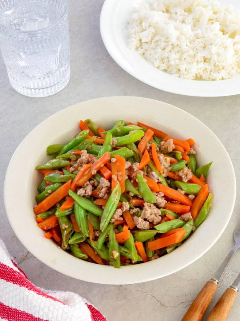 green bean stir-fry with carrots and minced pork on a white serving platter with a plate of steamed rice in the background.
