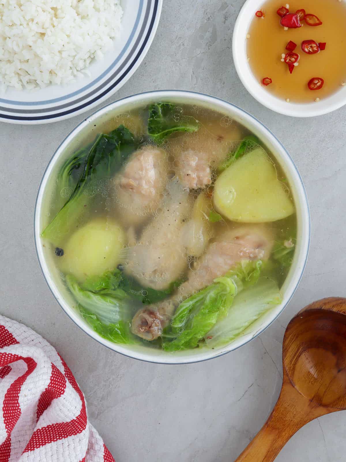 boiled chicken soup with potatoes and pechay in a white bowl with fish sauce in the background.