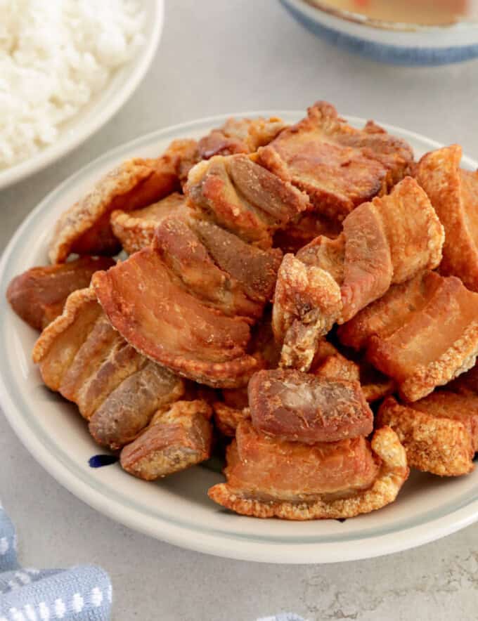 crispy pork belly in a white serving bowl with steamed rice and vinegar dipping sauce in the background.