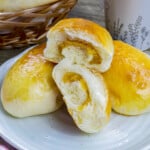 Filipino custard buns on a white plate with a basket of bread rolls and cup of coffee in the background.