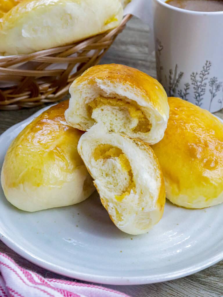 Filipino custard buns on a white plate with a basket of bread rolls and cup of coffee in the background.