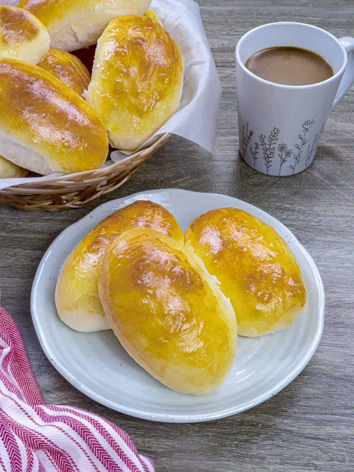 Yema Bread Rolls on a white plate with a cup of coffee and a basket of rolls in the background.