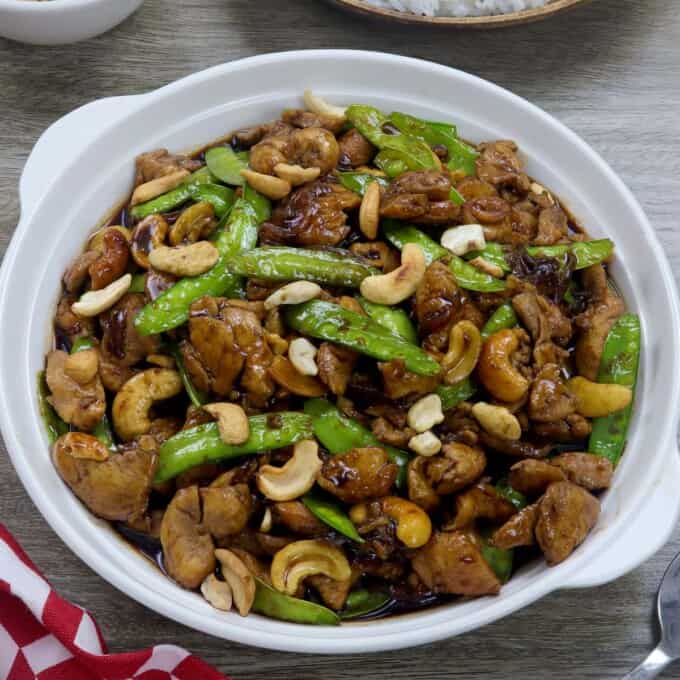 Cashew Chicken with Snow Peas in a white serving bowl with a plate of steamed rice and a small white bowl of cashews inn the background.