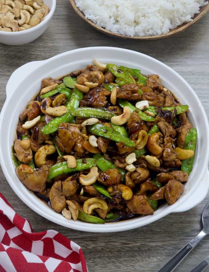 Cashew Chicken with Snow Peas in a white serving bowl with a plate of steamed rice and a small white bowl of cashews inn the background.