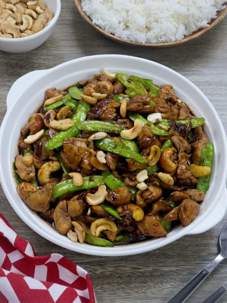 Cashew Chicken with Snow Peas in a white serving bowl with a plate of steamed rice and a small white bowl of cashews inn the background.