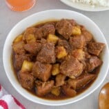 pork adobo with pineapple in a white serving bowl with a plate of steamed rice in the backround.